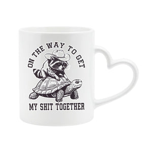 White heart-handle mug with a raccoon in a cowboy hat riding a tortoise and “On the way to get my shit together”