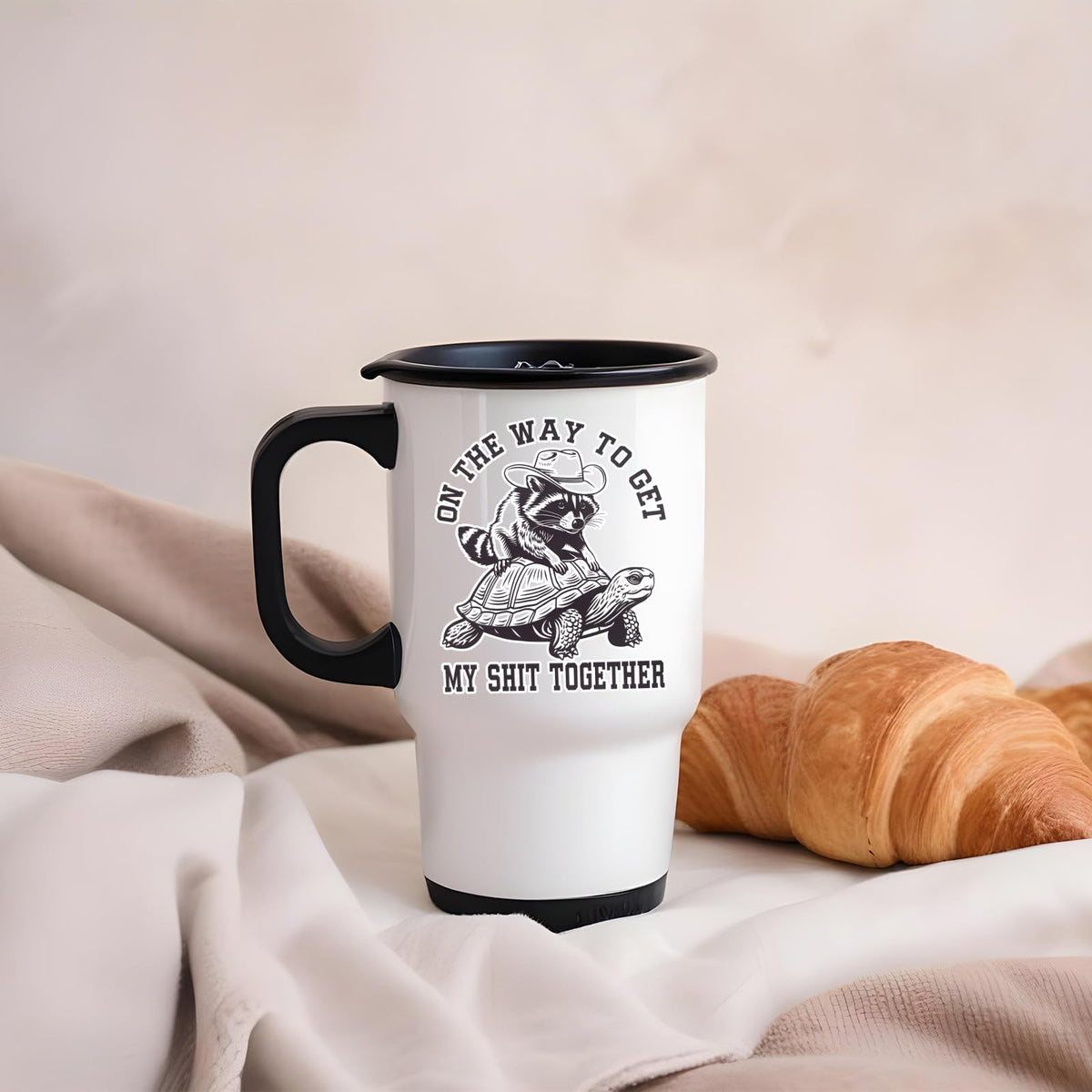 raccoon and tortoise travel mug on a soft blanket beside a croissant