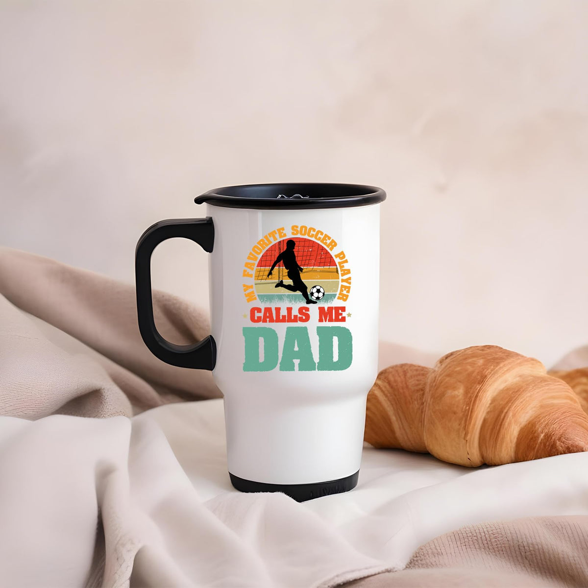 Soccer Dad travel mug on a soft blanket beside a croissant