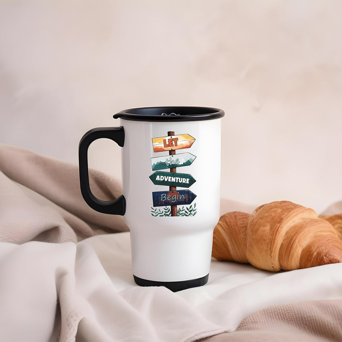 Let the Adventure Begin travel mug on a soft blanket beside a croissant