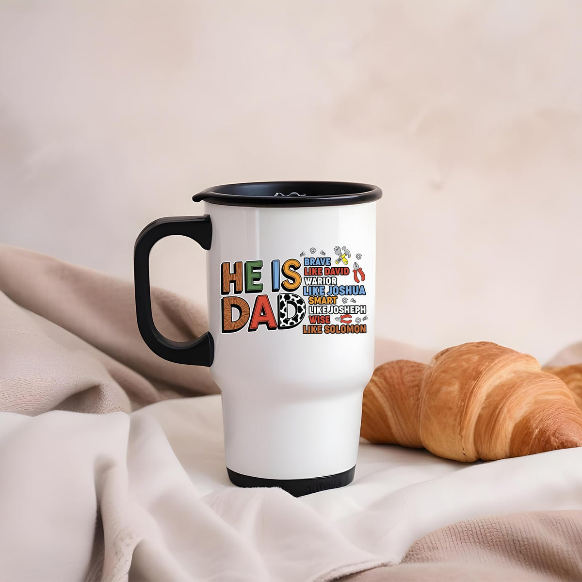 Travel mug with the He Is Dad Bible verse design on a blanket next to a croissant
