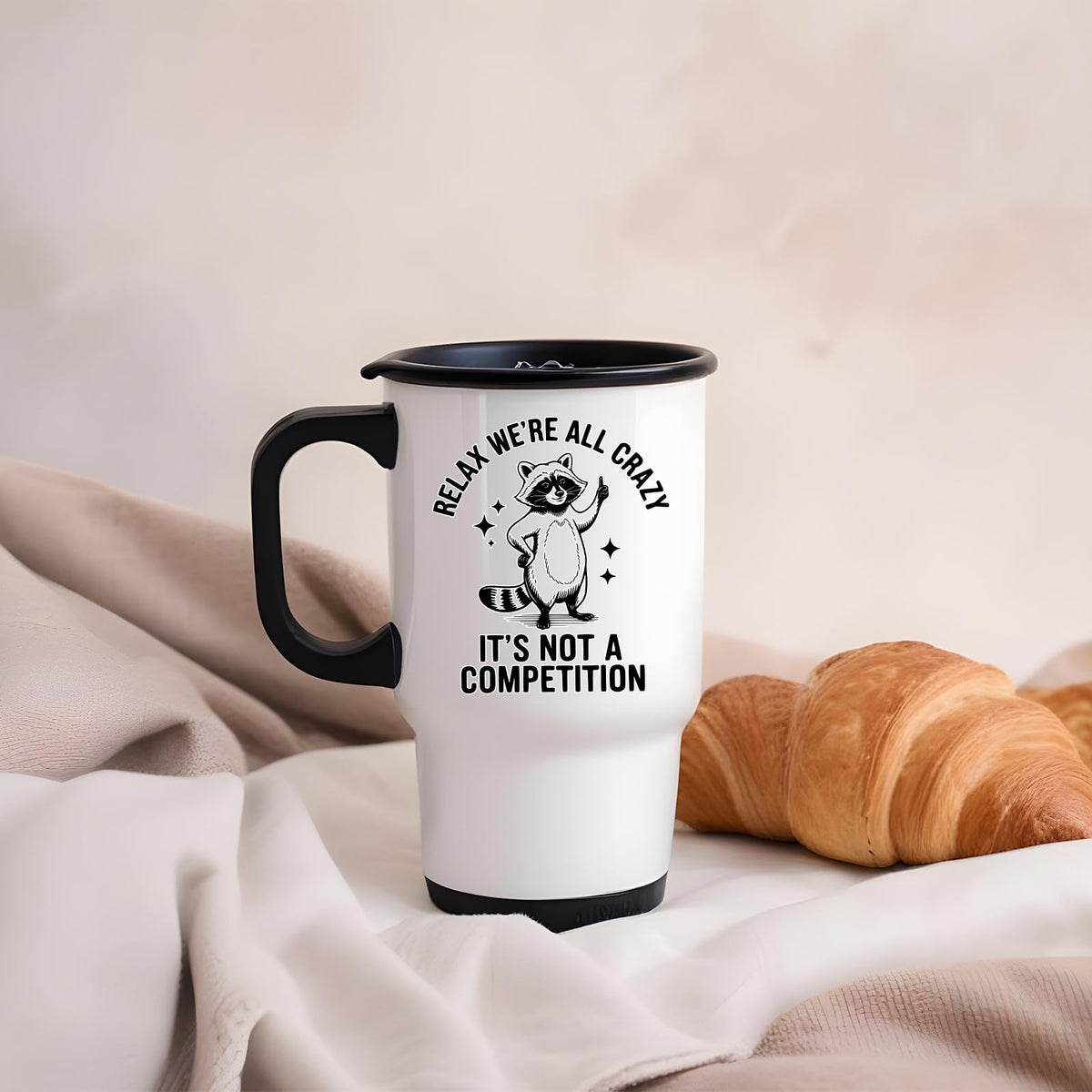 Relax We’re All Crazy raccoon travel mug on a soft blanket beside a croissant