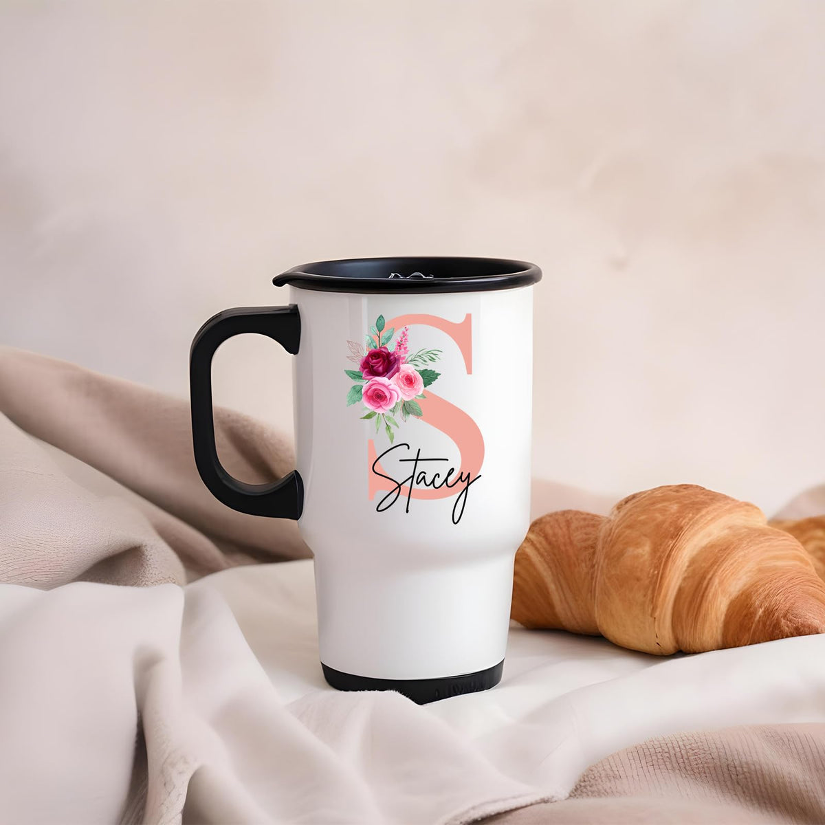 Travel mug with a blush pink initial with roses and an example name on a blanket next to a croissant
