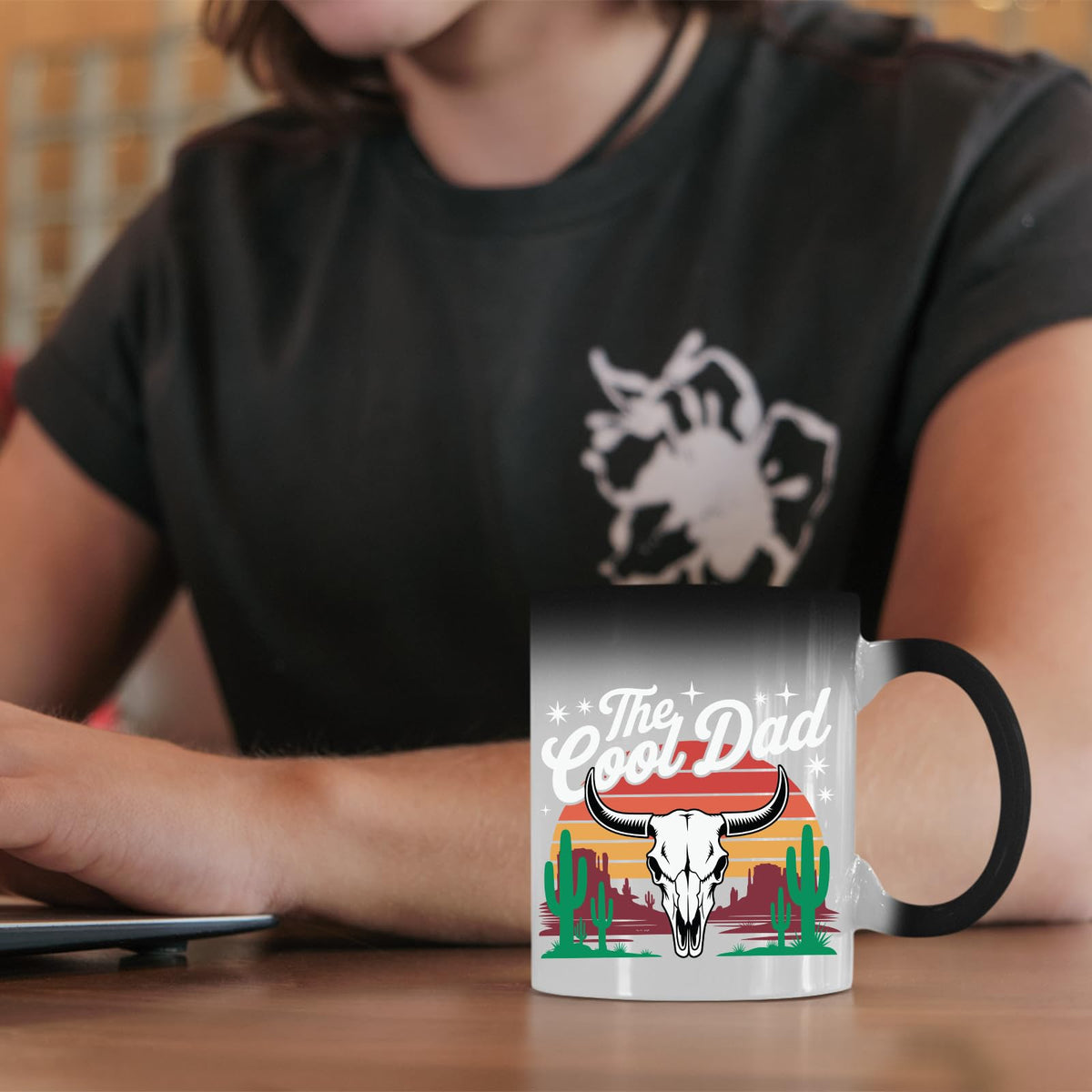 The Cool Dad bull skull colour changing mug on a desk beside a laptop