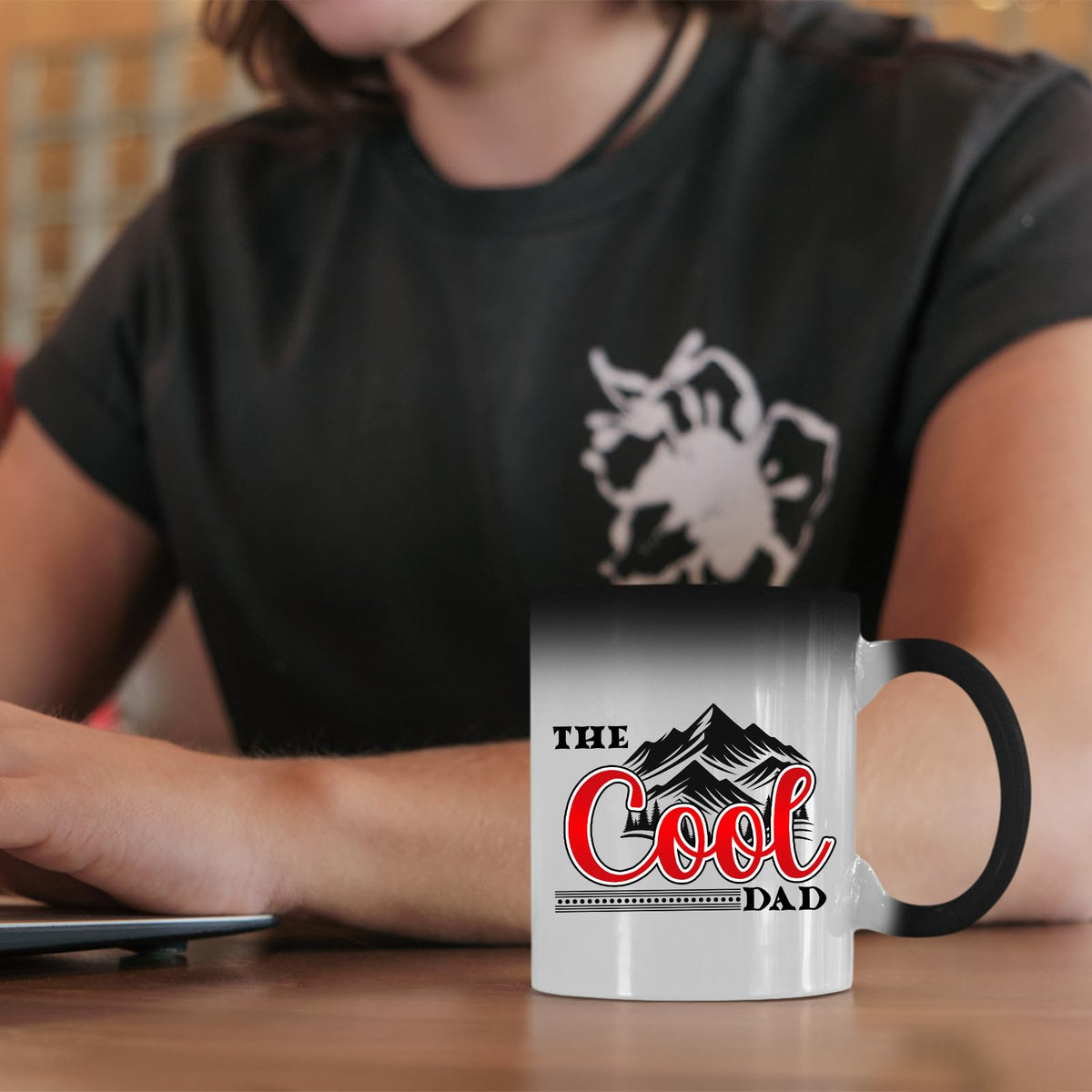 The Cool Dad mountains colour changing mug on a desk beside a laptop