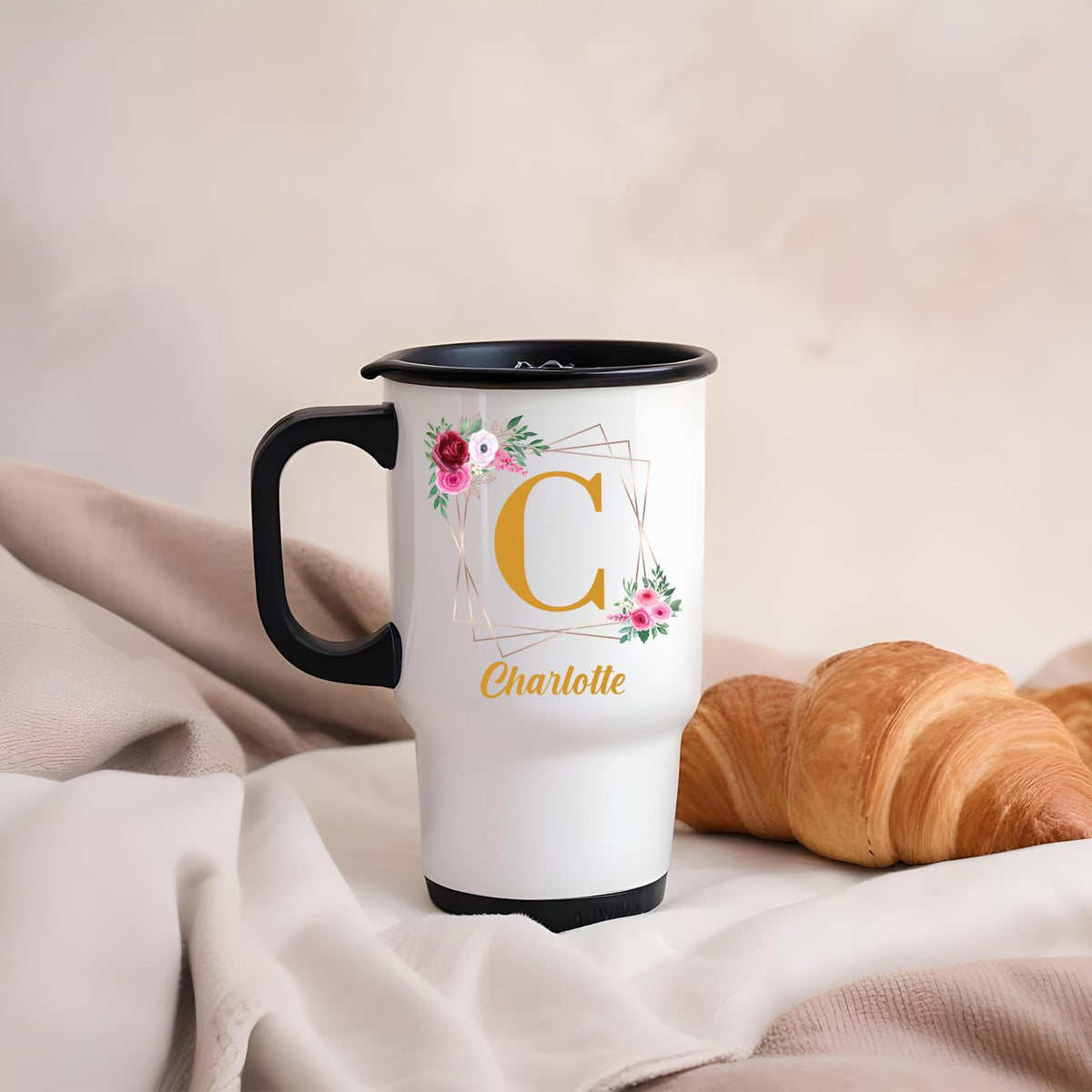 Travel mug with a gold initial in a floral frame and an example name on a blanket next to a croissant