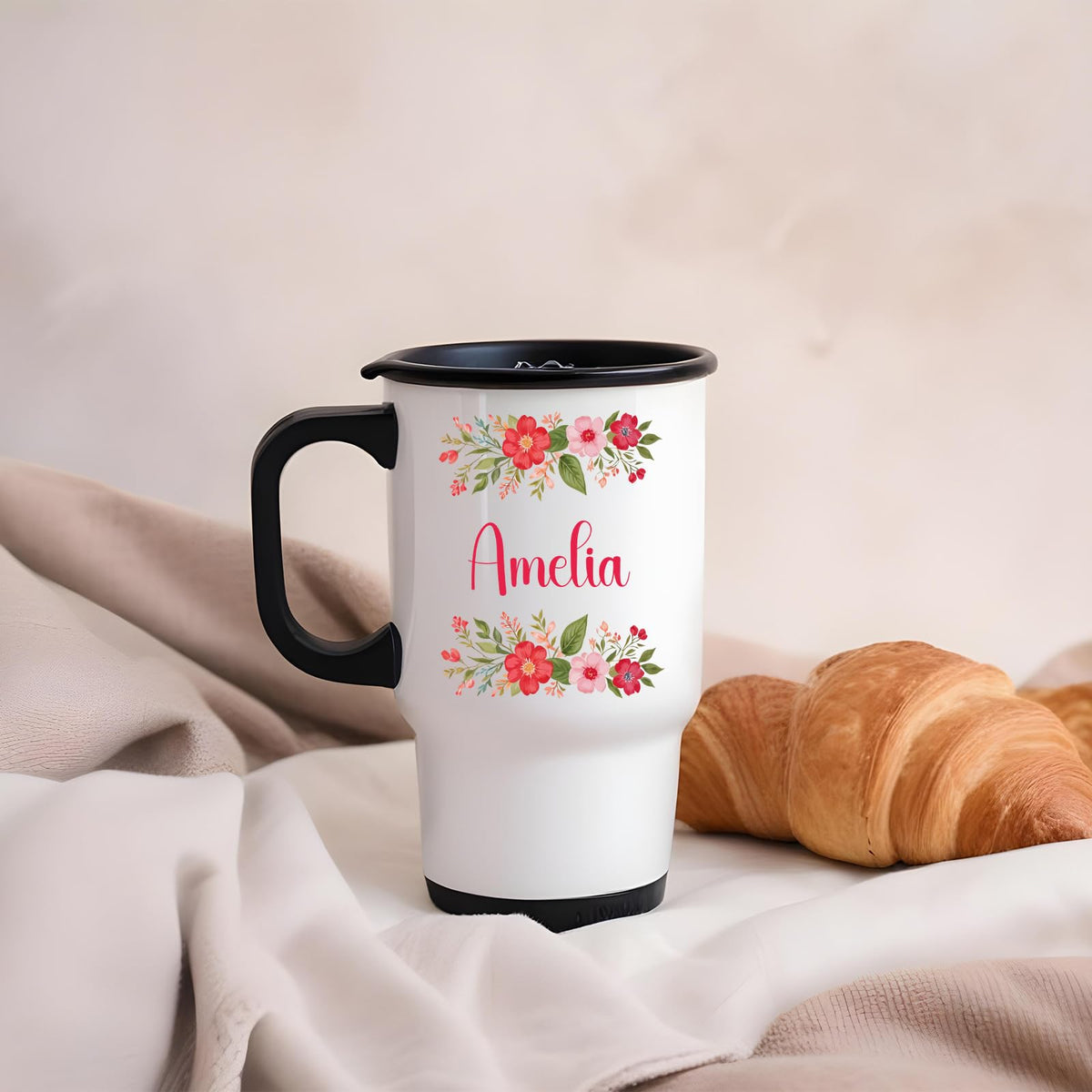 Travel mug with a red flower border around an example name on a blanket next to a croissant