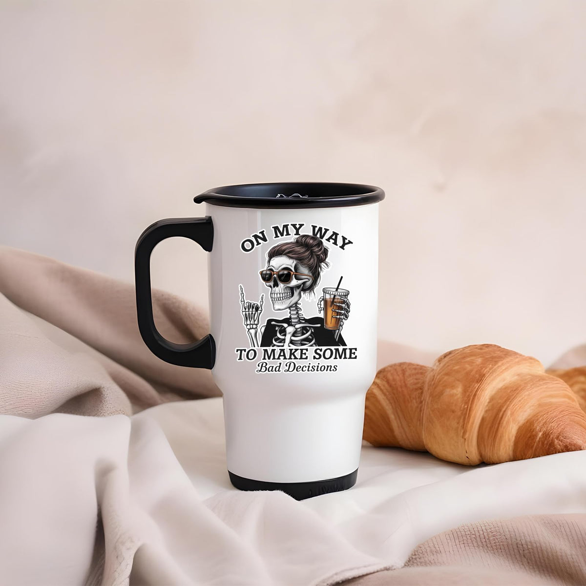 Bad Decisions skeleton travel mug on a soft blanket beside a croissant