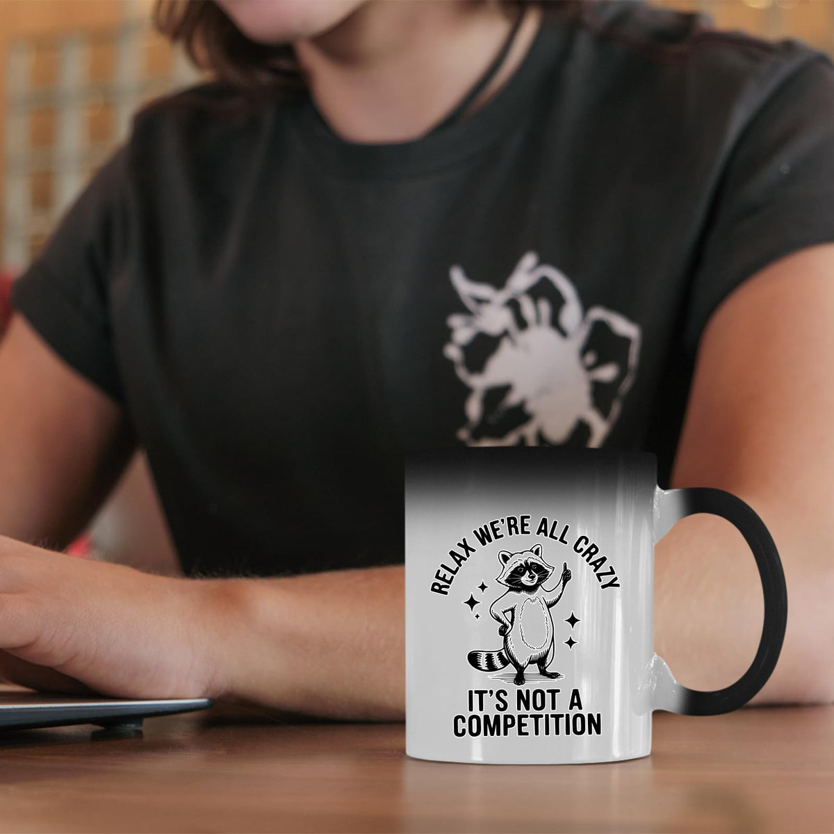 Relax We’re All Crazy raccoon colour changing mug on a desk beside a laptop