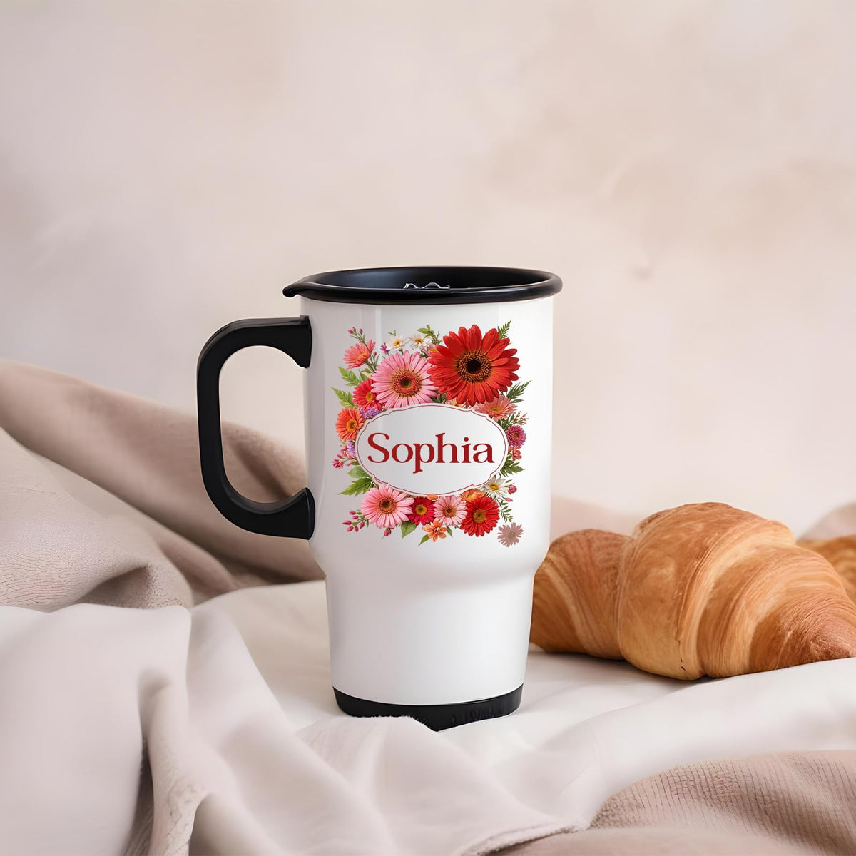 Travel mug with a red gerbera wreath around an example name beside a croissant