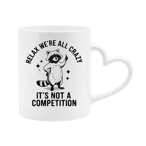 White heart-handle mug with a raccoon and “Relax, we’re all crazy – it’s not a competition”