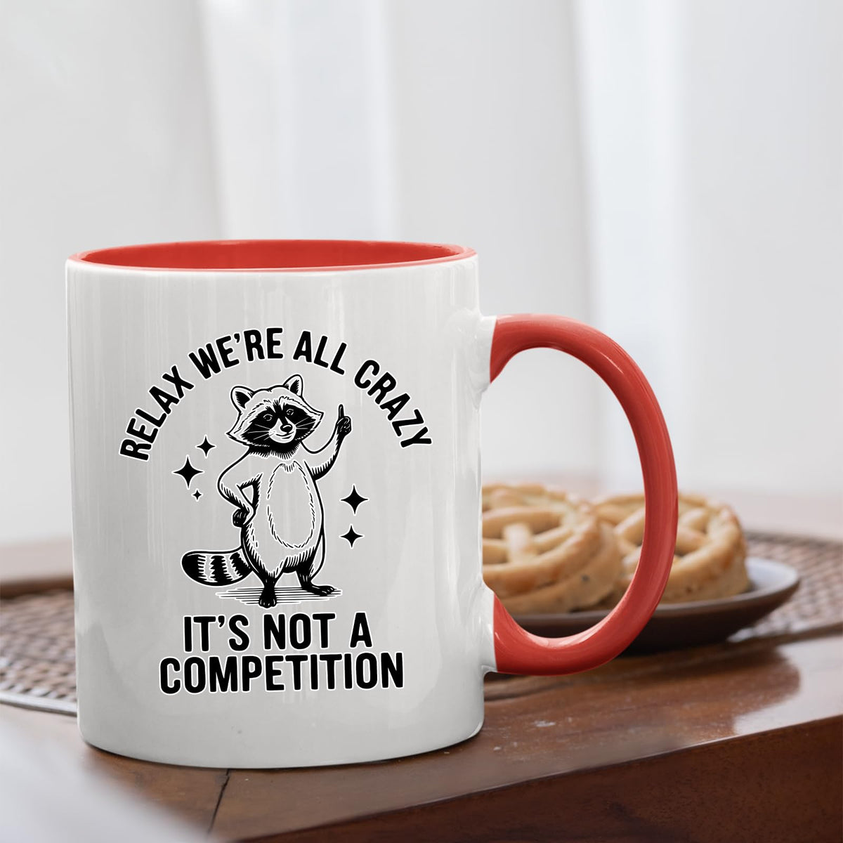 Relax We’re All Crazy raccoon mug with a red handle beside biscuits