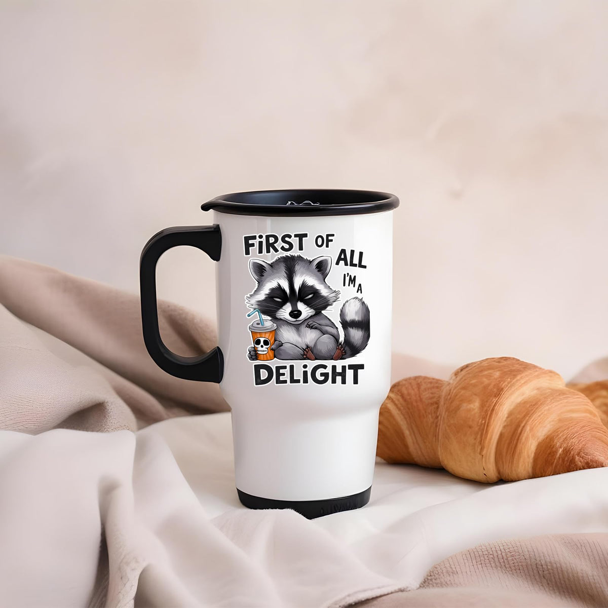 First of All I’m a Delight raccoon travel mug on a soft blanket beside a croissant