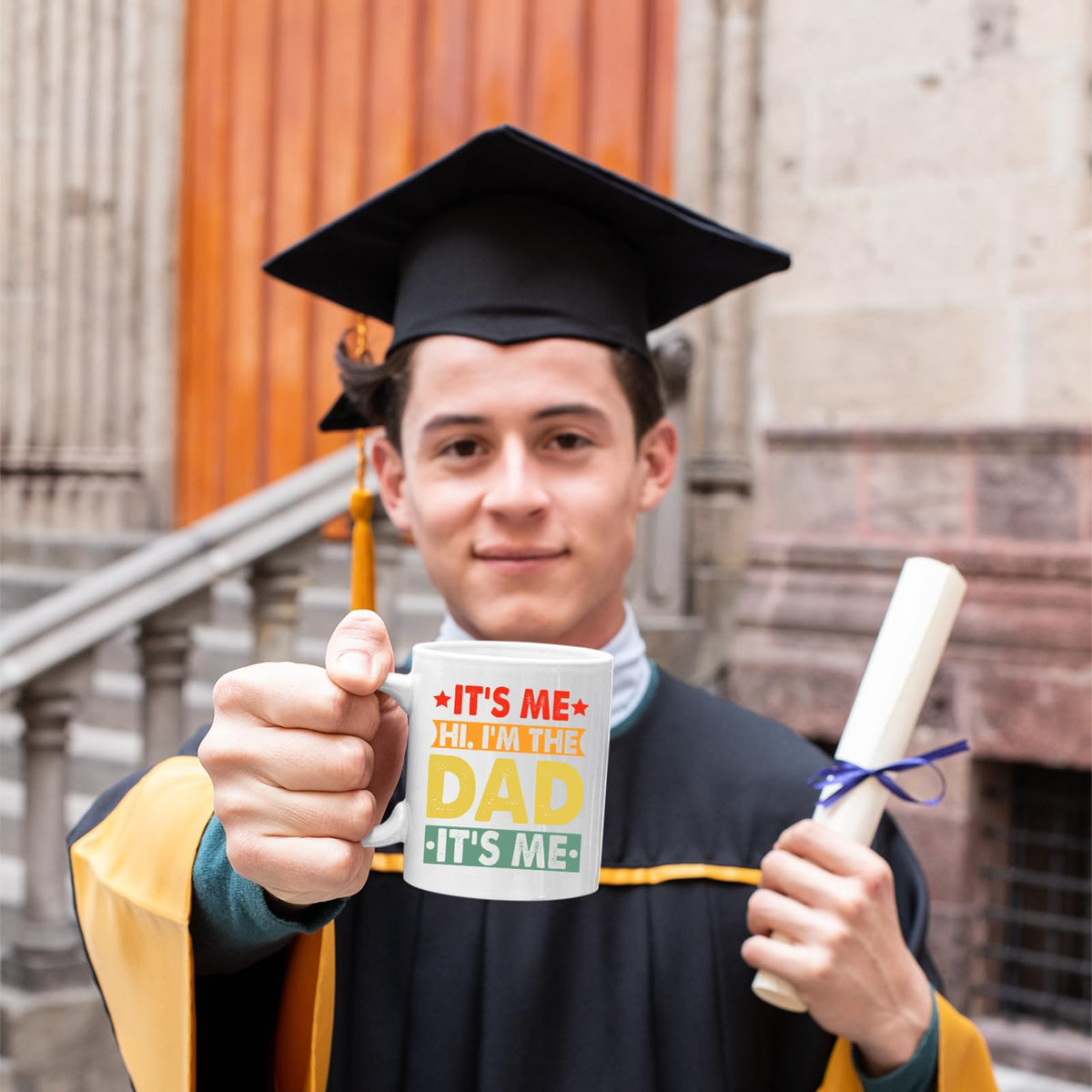 Graduate in cap and gown holding the It’s Me Hi I’m the Dad mug