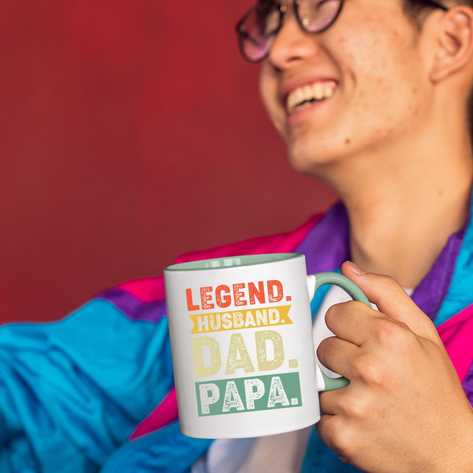 Smiling person in a colourful jacket holding the Legend Husband Dad Papa mug with a light green handle