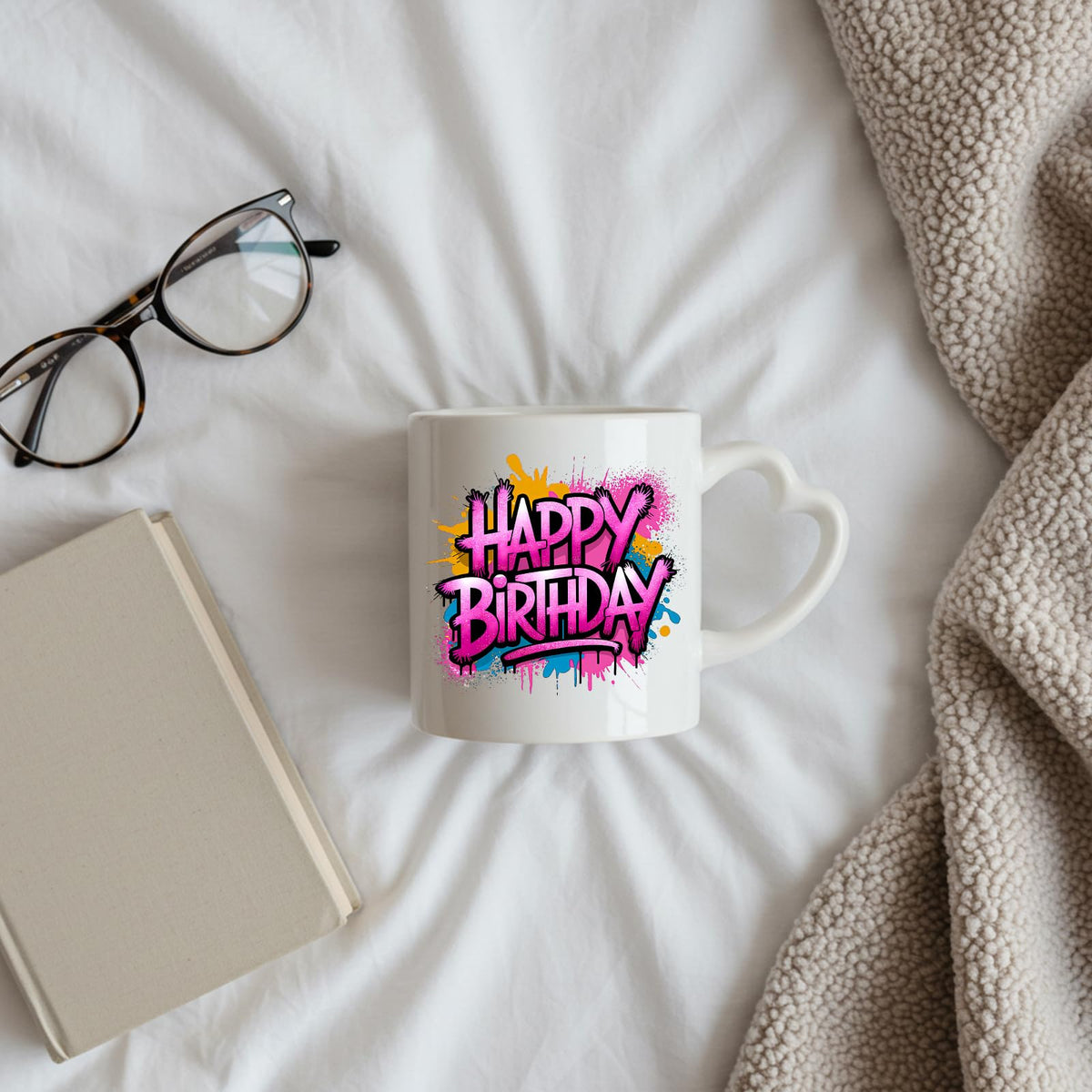 Happy Birthday pink graffiti splash heart-handle mug on a bed beside glasses and a book