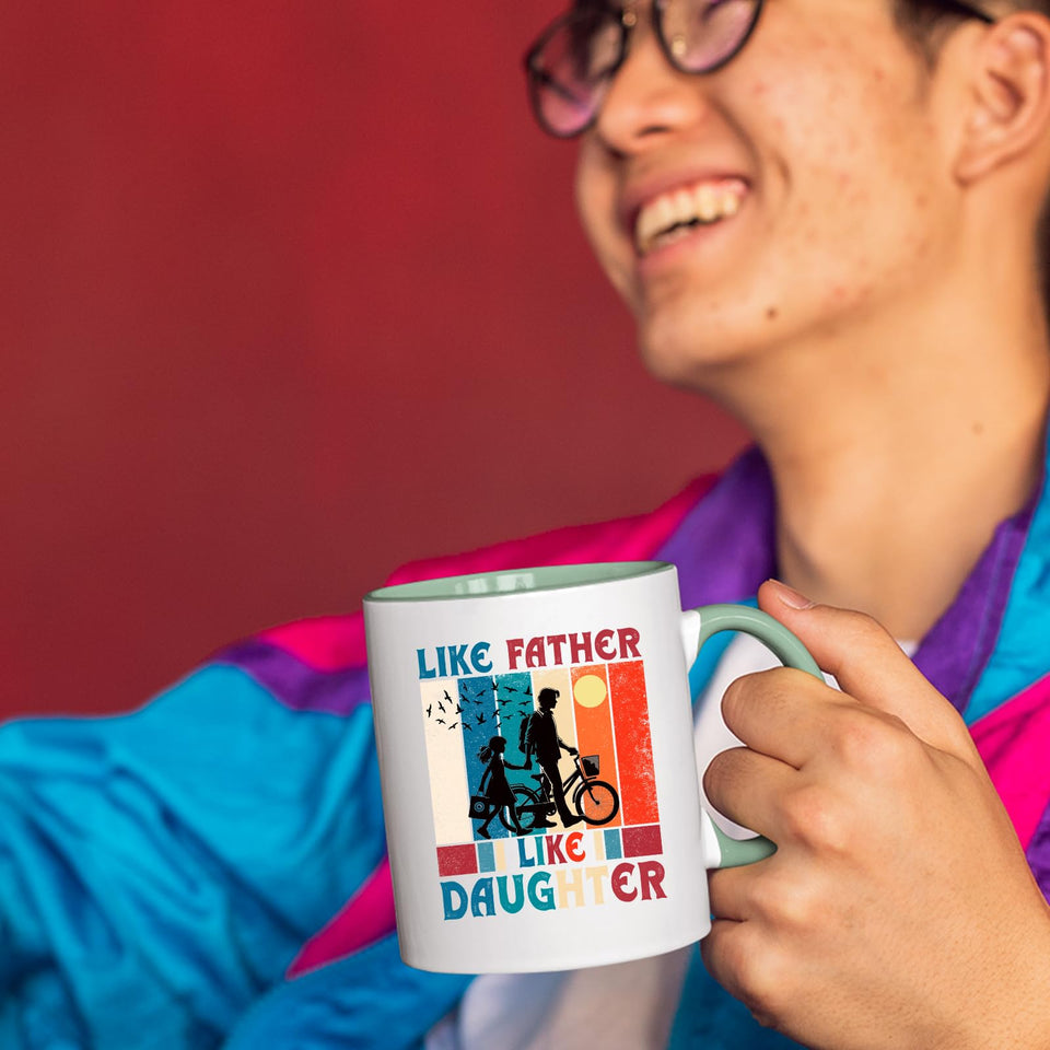 Smiling person in a colourful jacket holding the Like Father Like Daughter mug with a light green handle