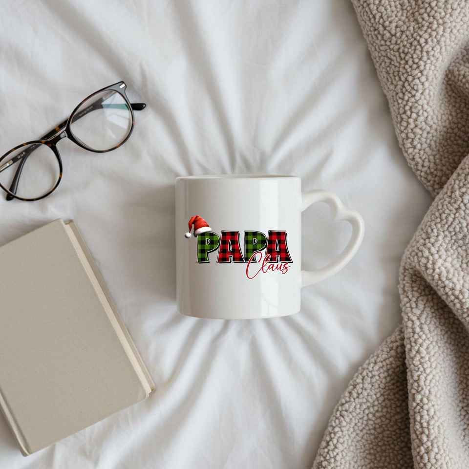 Papa Claus heart-handle mug on a bed beside glasses and a book