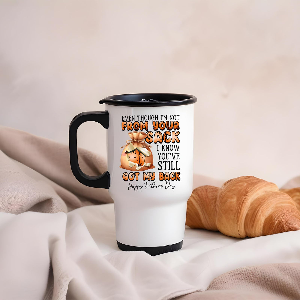 Not From Your Sack travel mug on a soft blanket beside a croissant