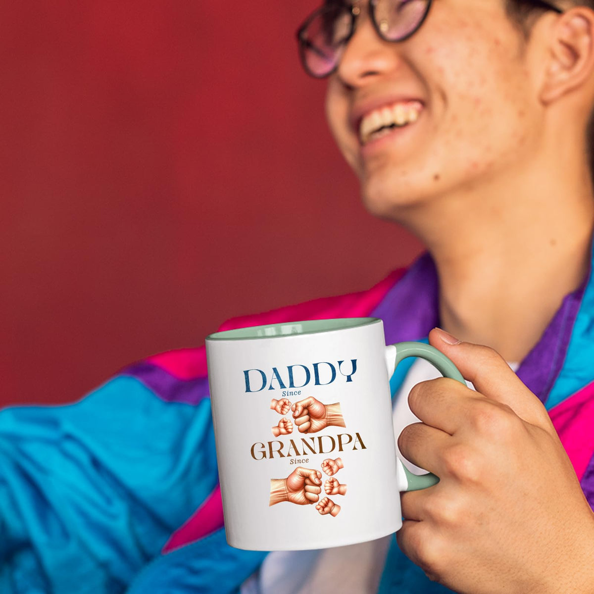 Smiling person in a colourful jacket holding the Daddy and Grandpa fist bump mug with a light green handle