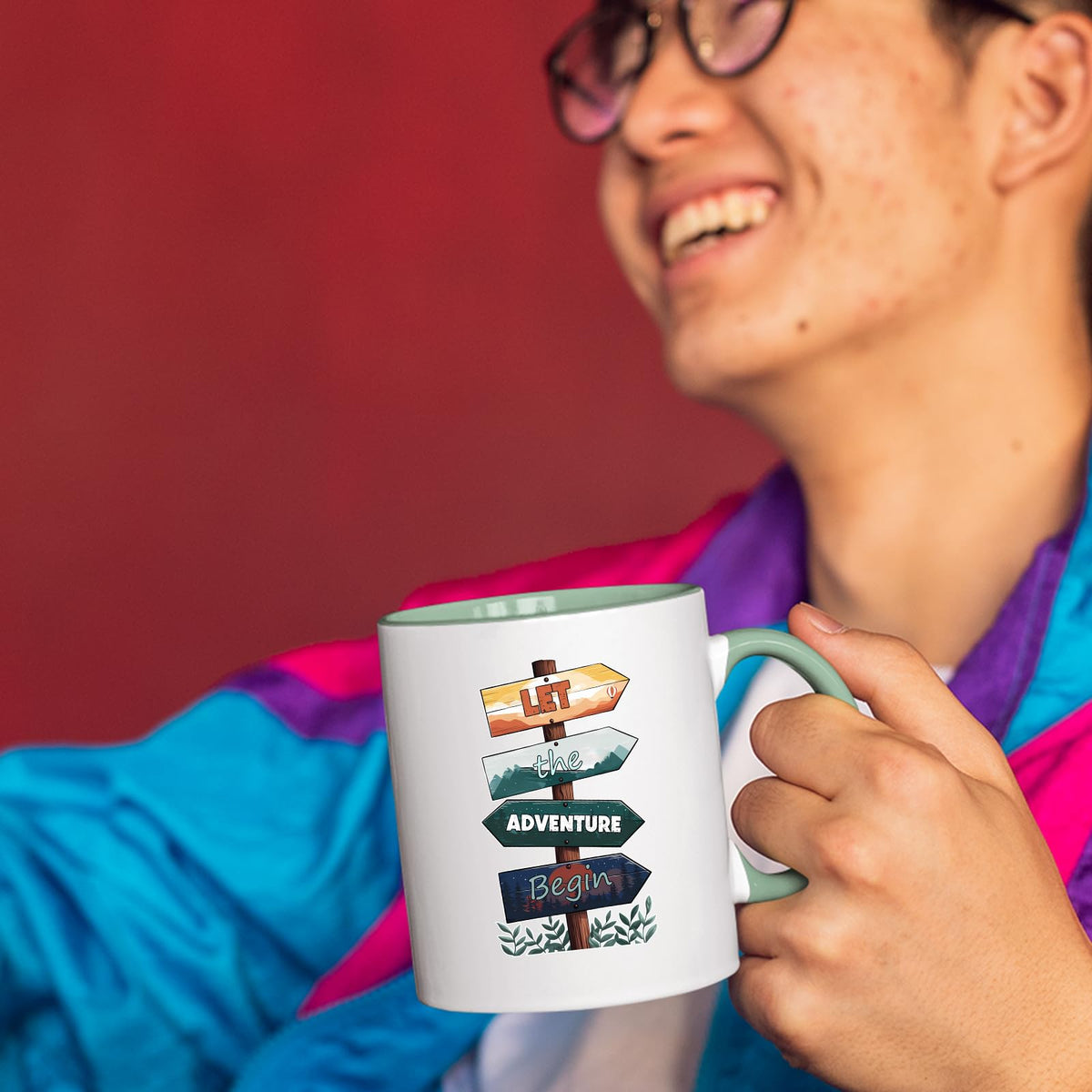 Smiling person in a colourful jacket holding the Let the Adventure Begin mug with a light green handle