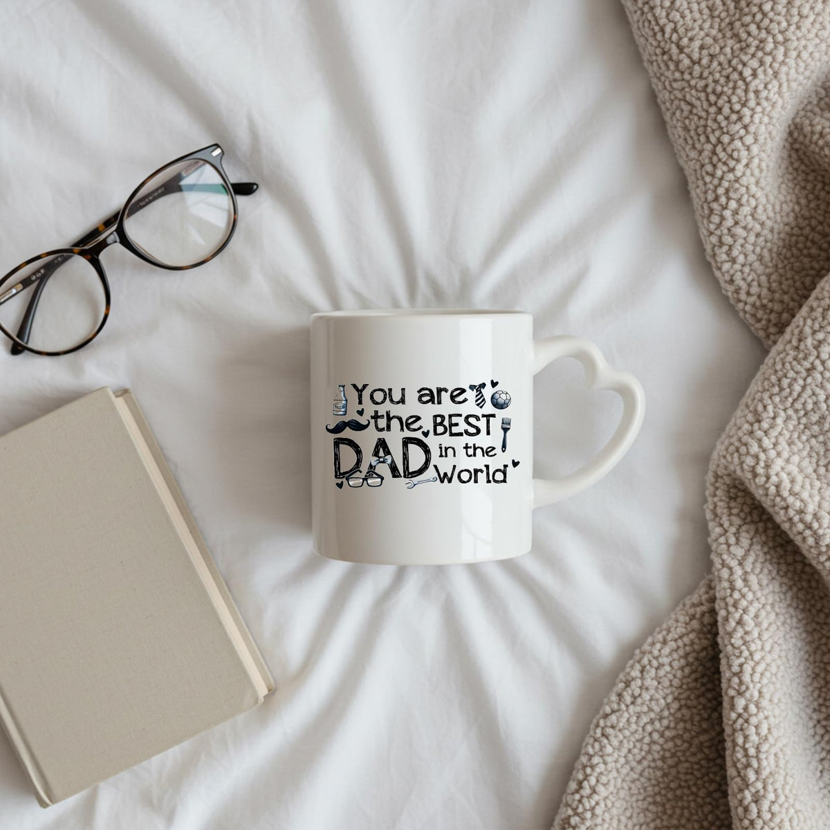 Best Dad in the World sketch heart-handle mug on a bed beside glasses and a book