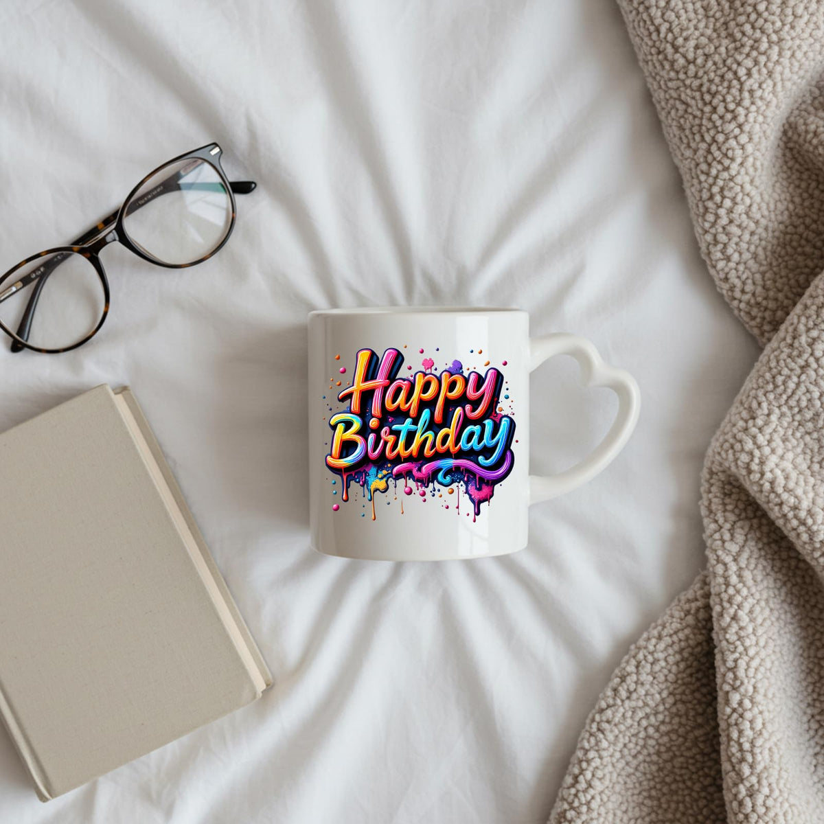 Happy Birthday rainbow drip heart-handle mug on a bed beside glasses and a book