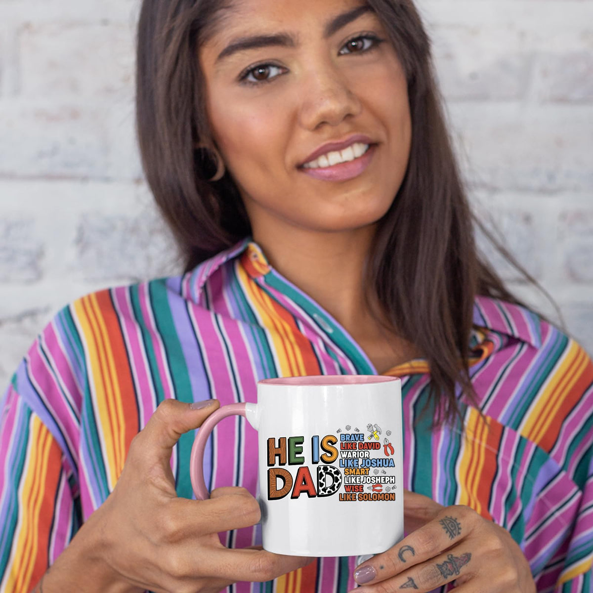 Woman in a striped shirt holding a colour handle mug with the He Is Dad Bible verse design