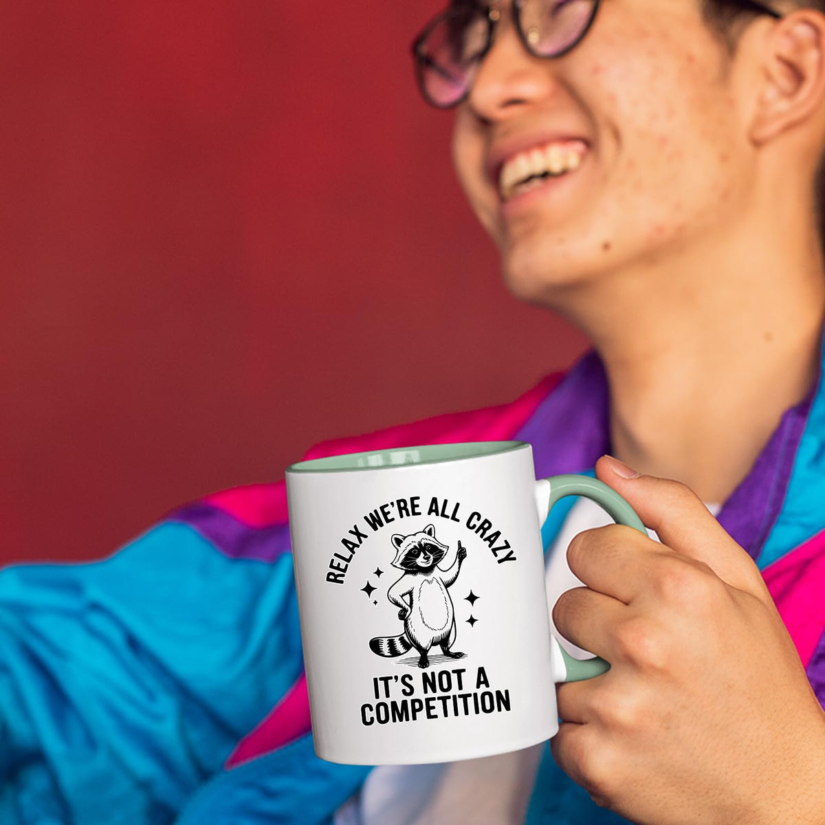 Smiling person in a colourful jacket holding the Relax We’re All Crazy raccoon mug with a light green handle