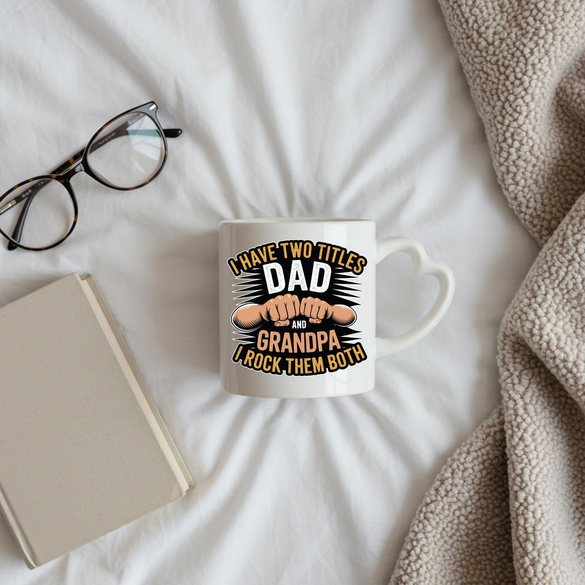 Two Titles Dad and Grandpa heart-handle mug on a bed beside glasses and a book