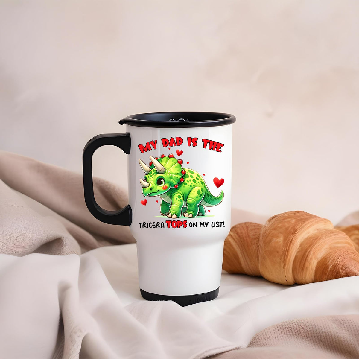 Triceratops dad travel mug on a soft blanket beside a croissant