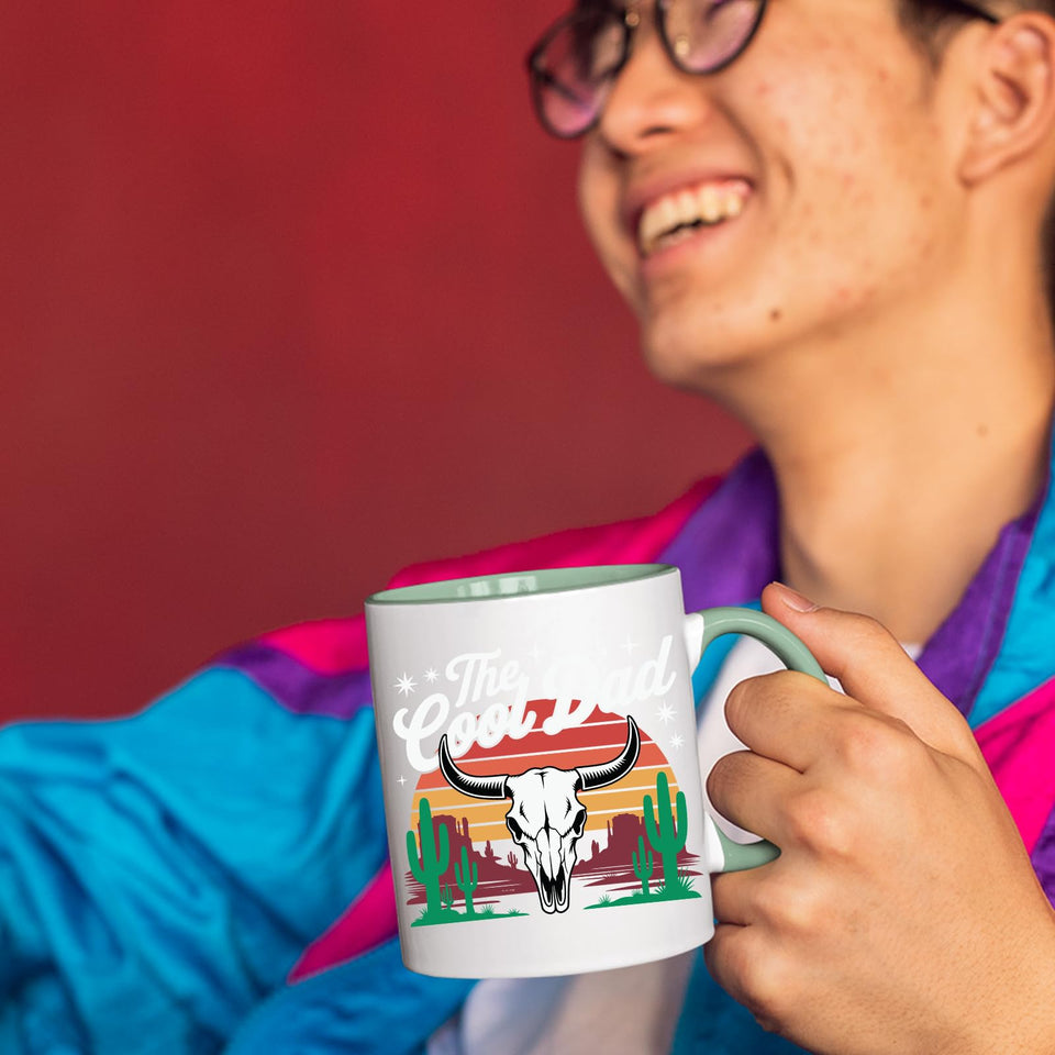 Smiling person in a colourful jacket holding The Cool Dad bull skull mug with a light green handle