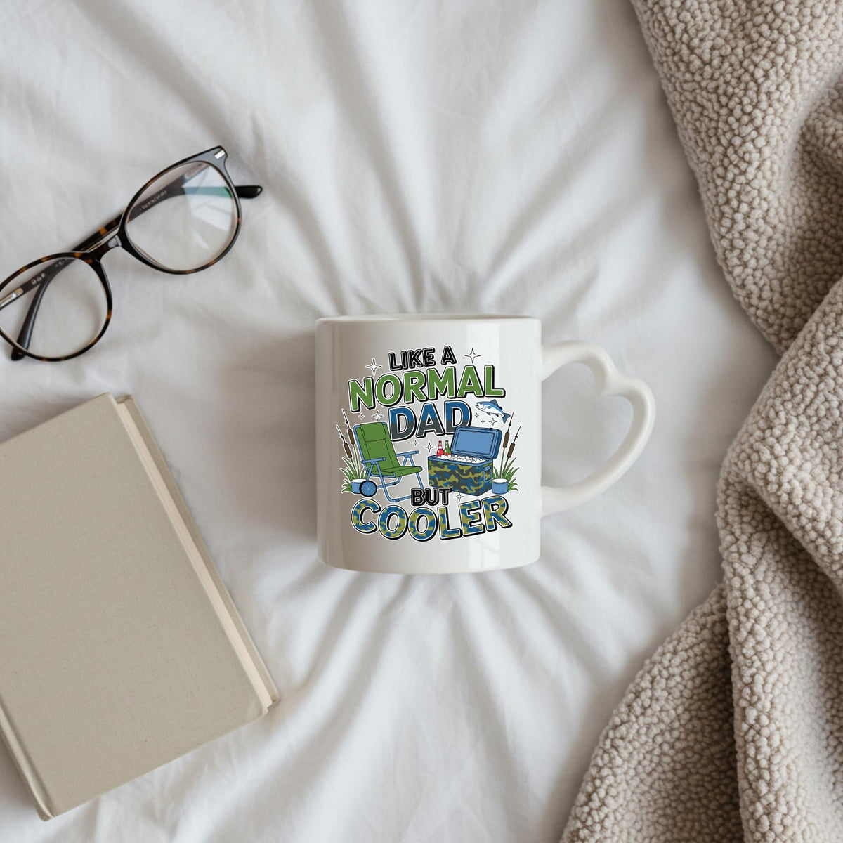 Like a Normal Dad But Cooler heart-handle mug on a bed beside glasses and a book