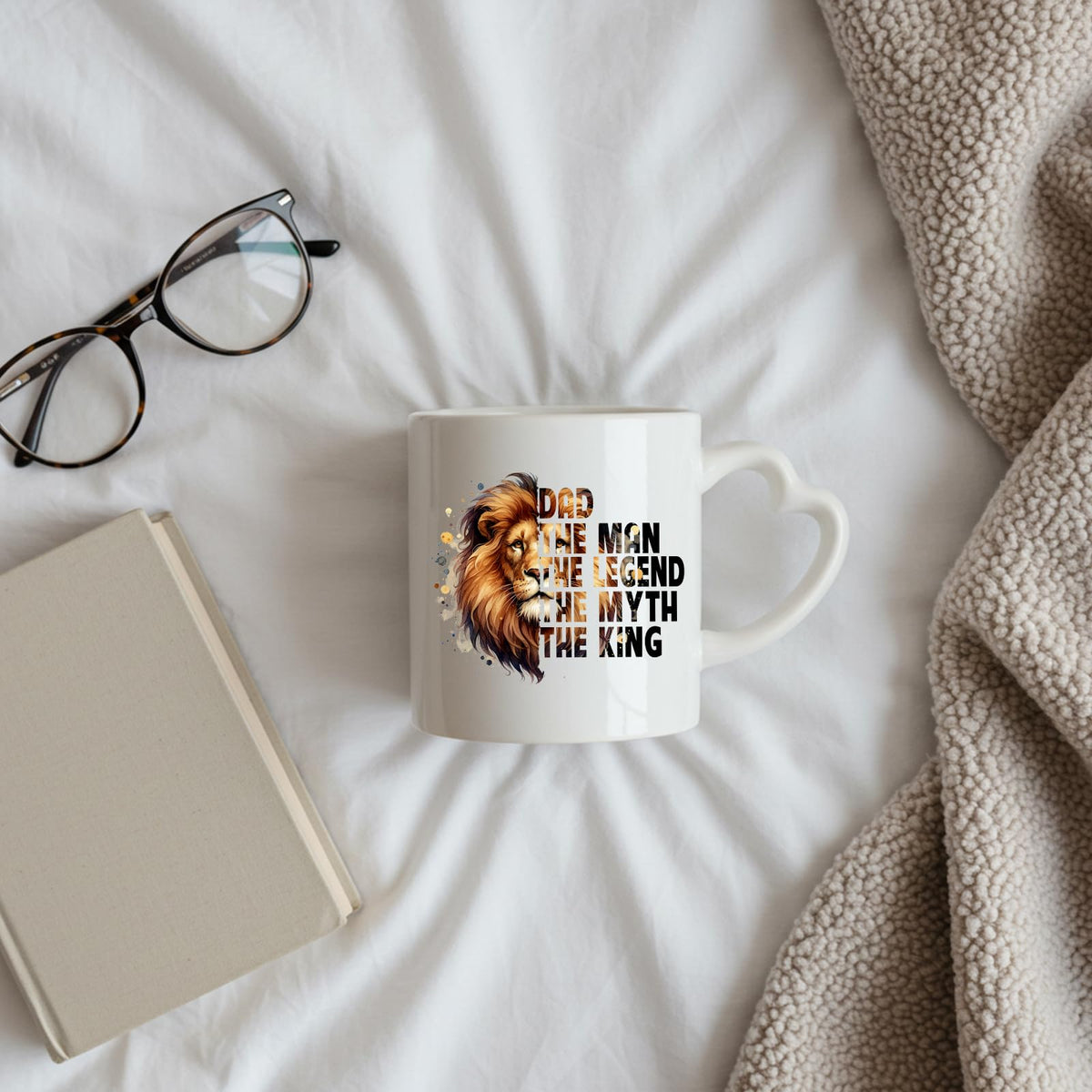Dad the King lion heart-handle mug on a bed beside glasses and a book