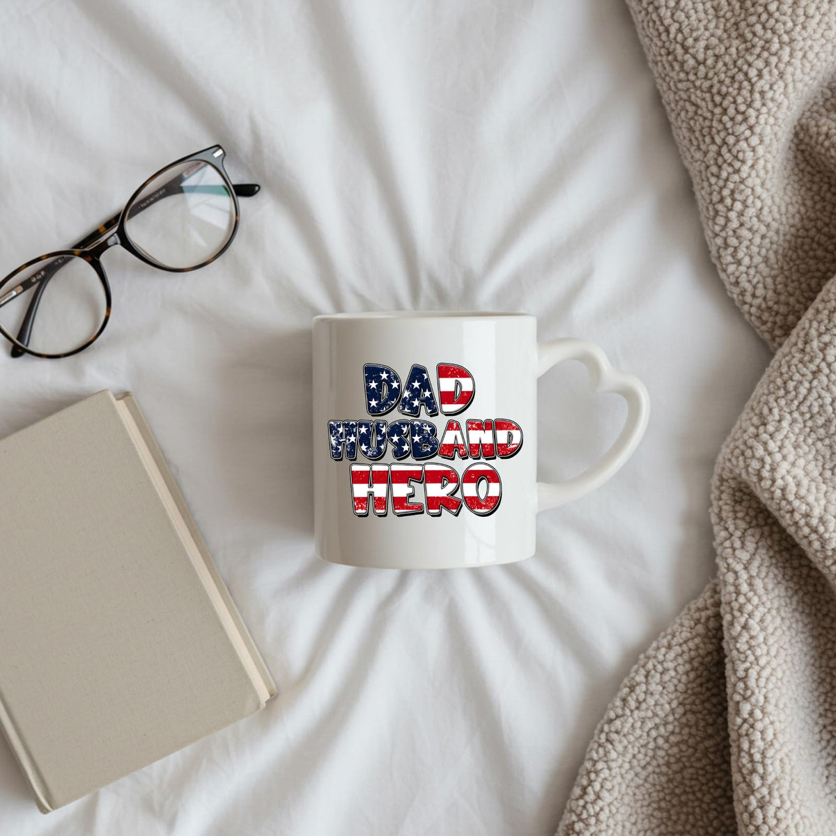Dad Husband Hero heart-handle mug on a bed beside glasses and a book