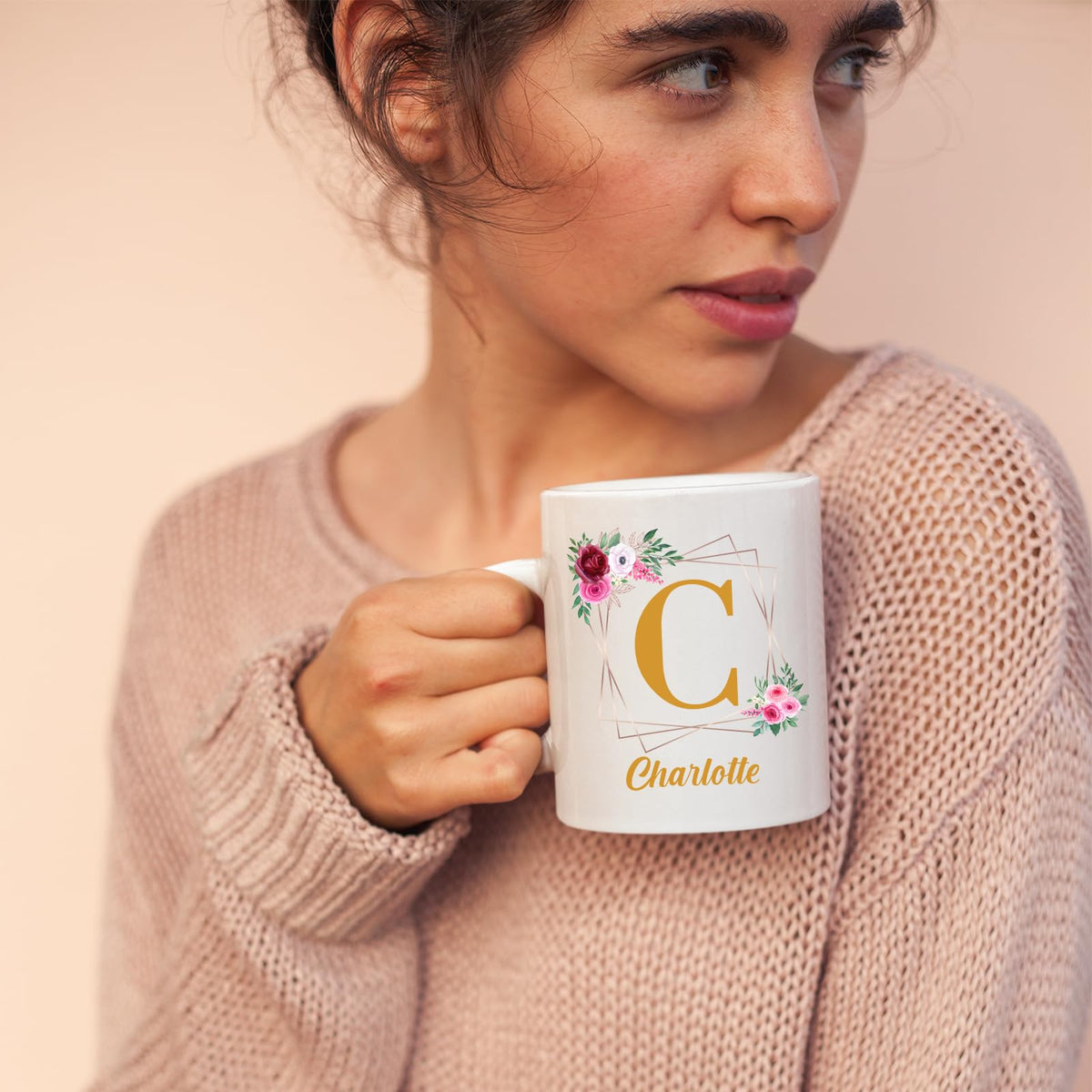 Woman in a pink jumper holding a mug with a gold initial in a floral frame and an example name