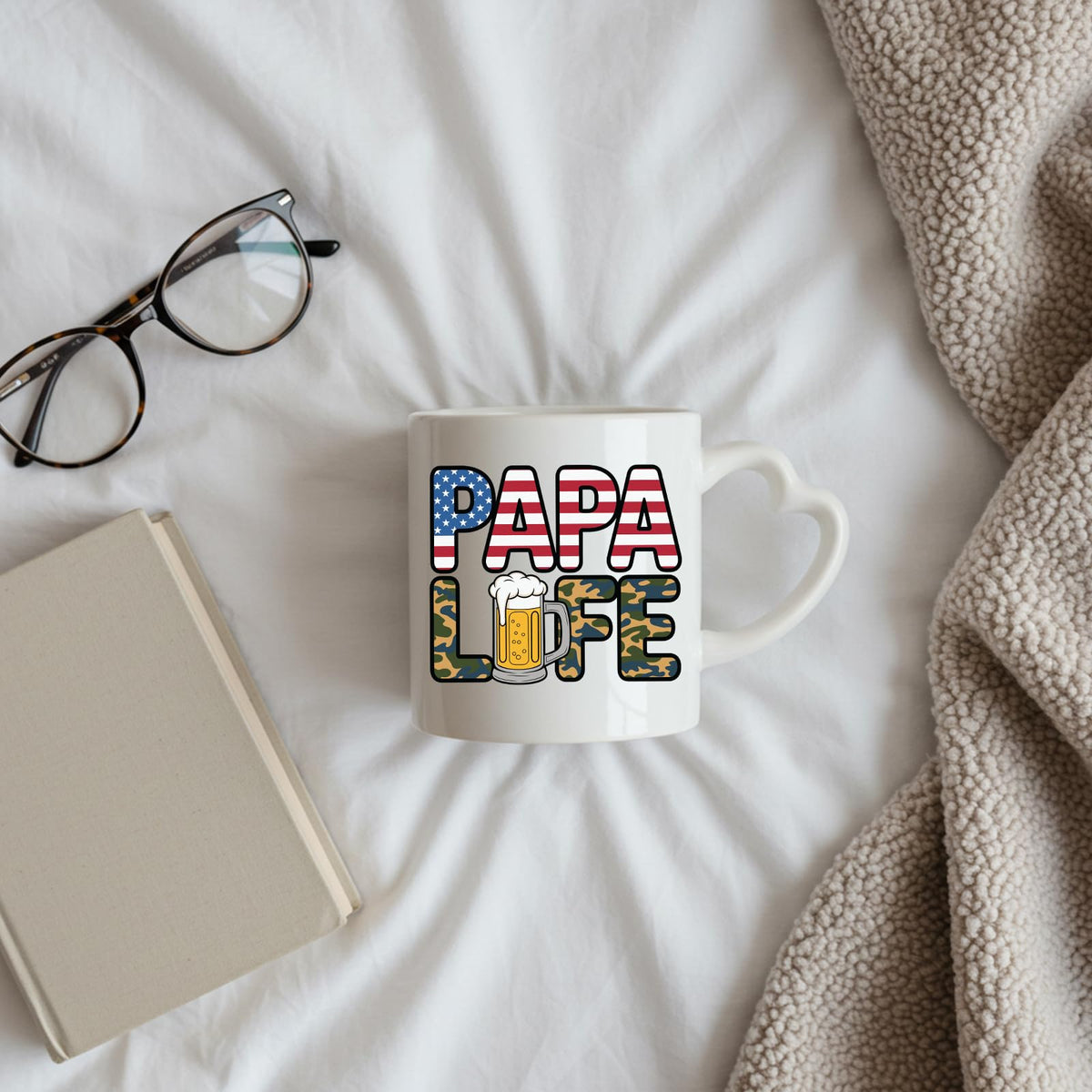 Papa Life heart-handle mug on a bed beside glasses and a book