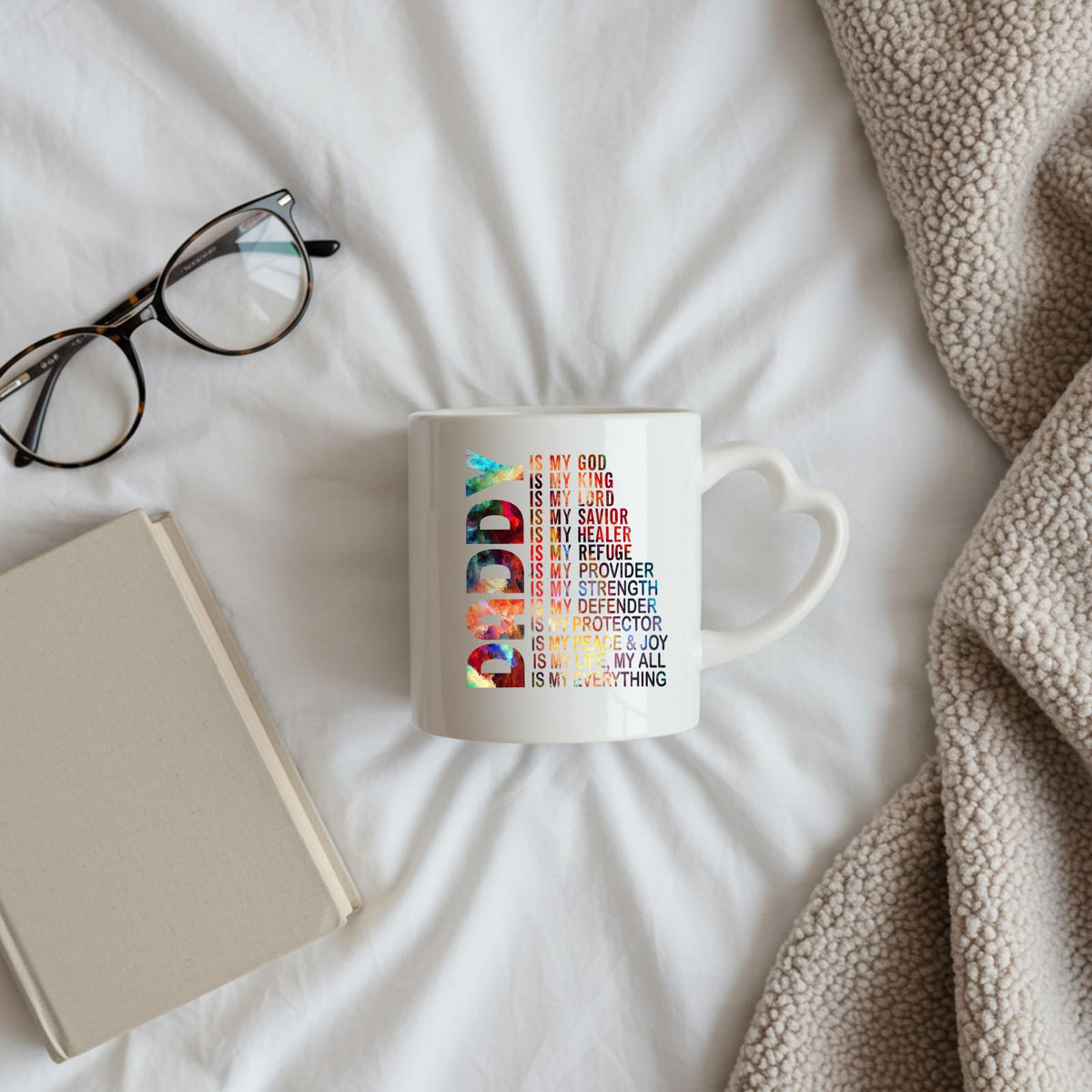 Daddy Is My God heart-handle mug on a bed beside glasses and a book