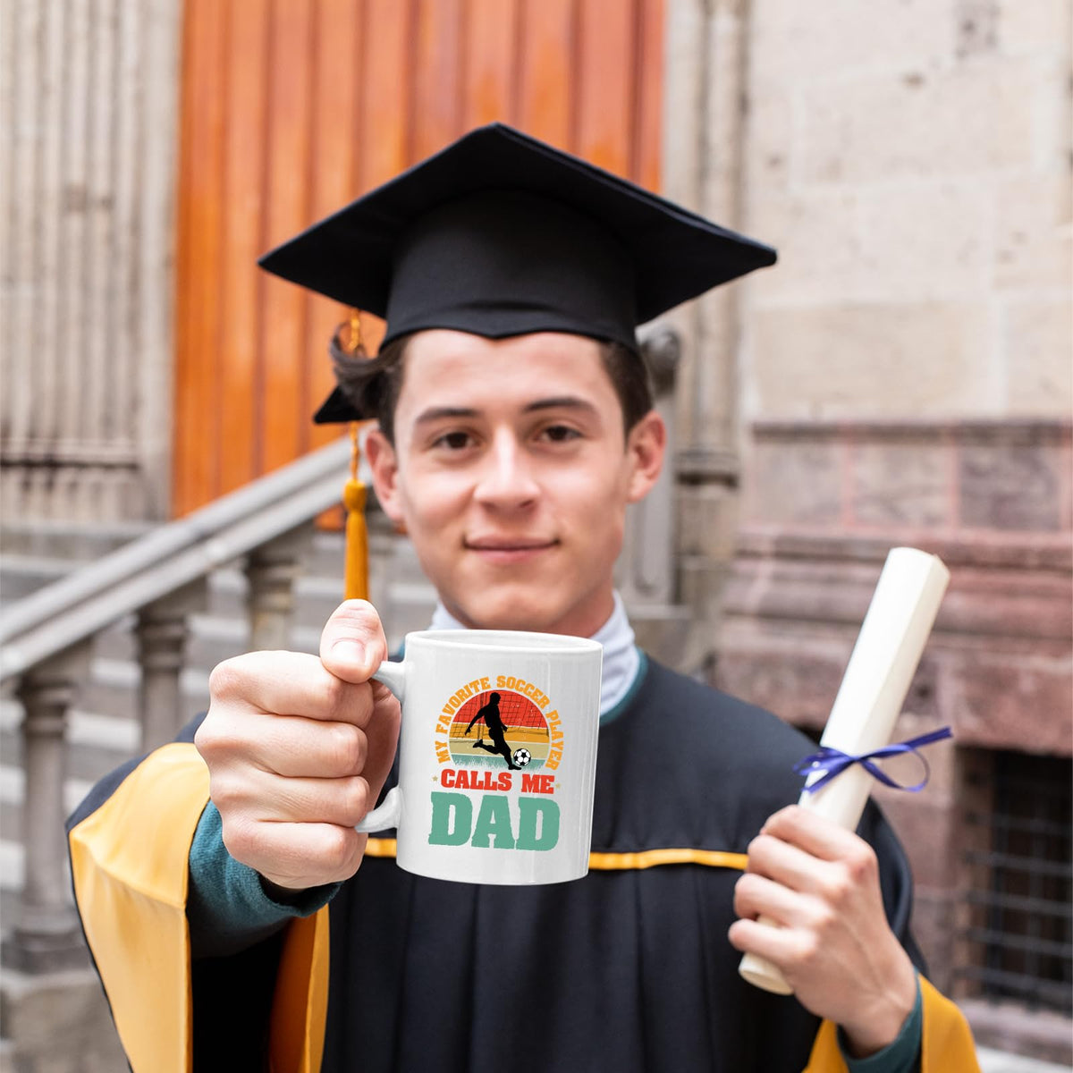 Graduate in cap and gown holding the Soccer Dad mug