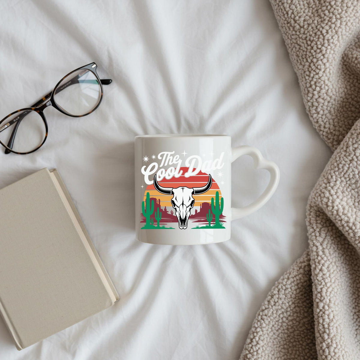 The Cool Dad bull skull heart-handle mug on a bed beside glasses and a book
