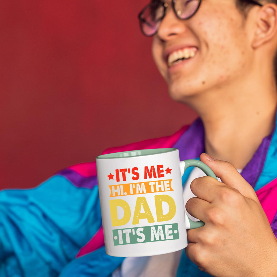 Smiling person in a colourful jacket holding the It’s Me Hi I’m the Dad mug with a light green handle
