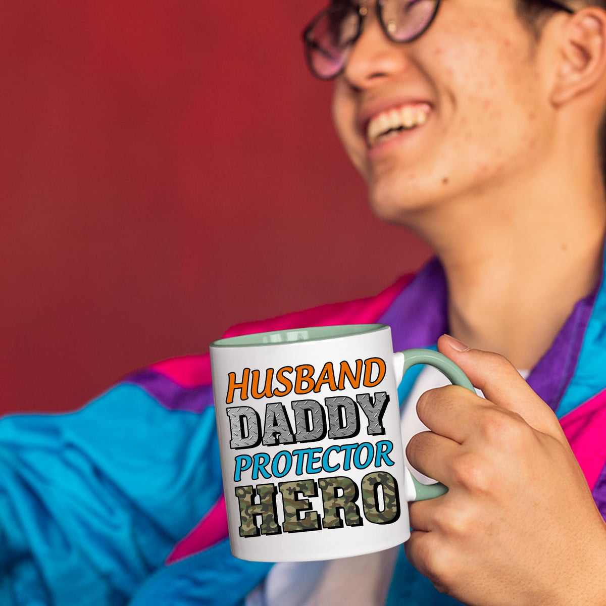 Smiling person in a colourful jacket holding the Husband Daddy Protector Hero camo mug with a light green handle
