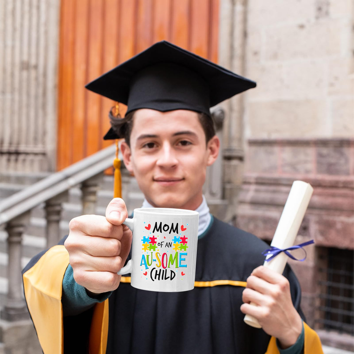 Graduate in cap and gown holding the Mom of an Au-Some Child mug
