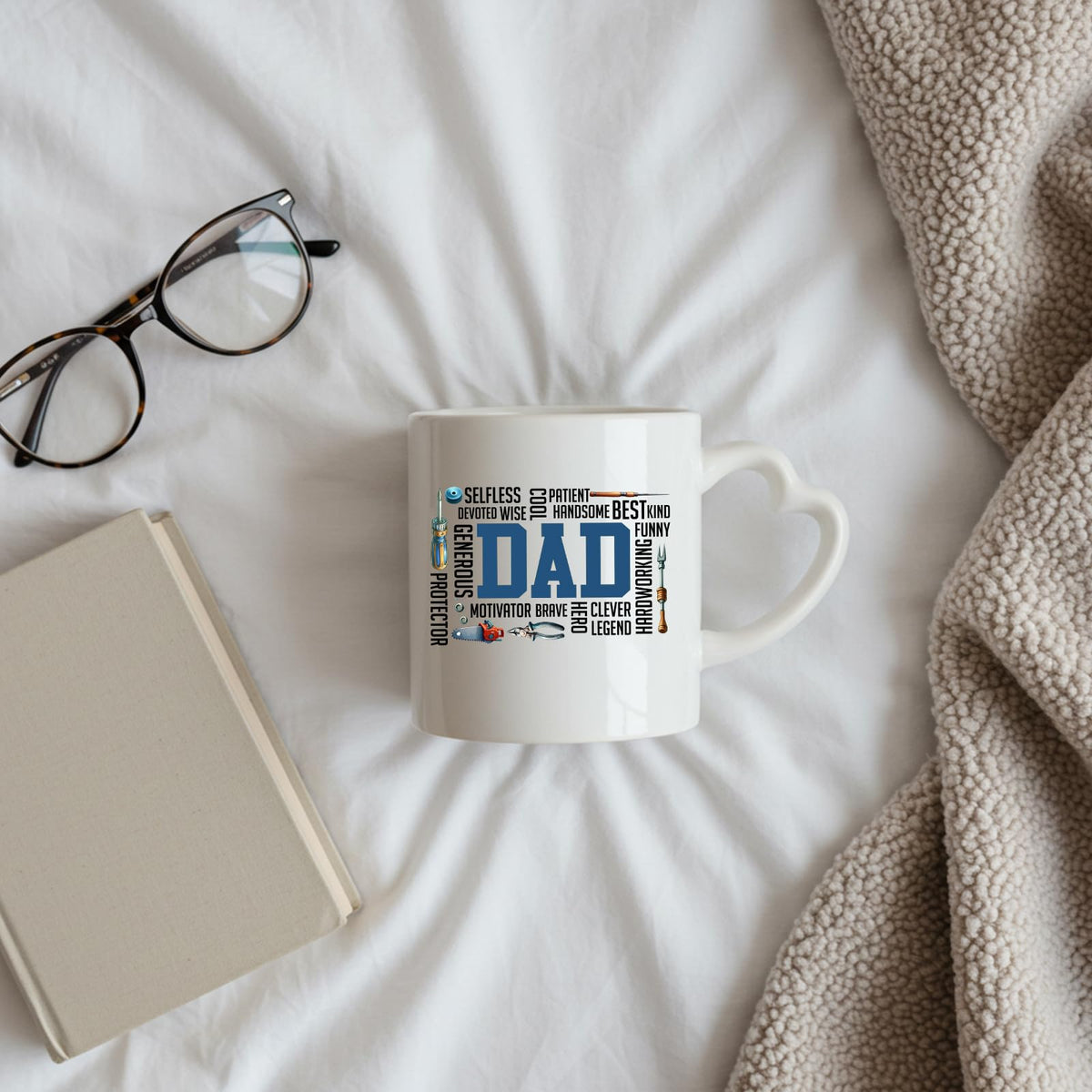 DAD word cloud heart-handle mug on a bed beside glasses and a book