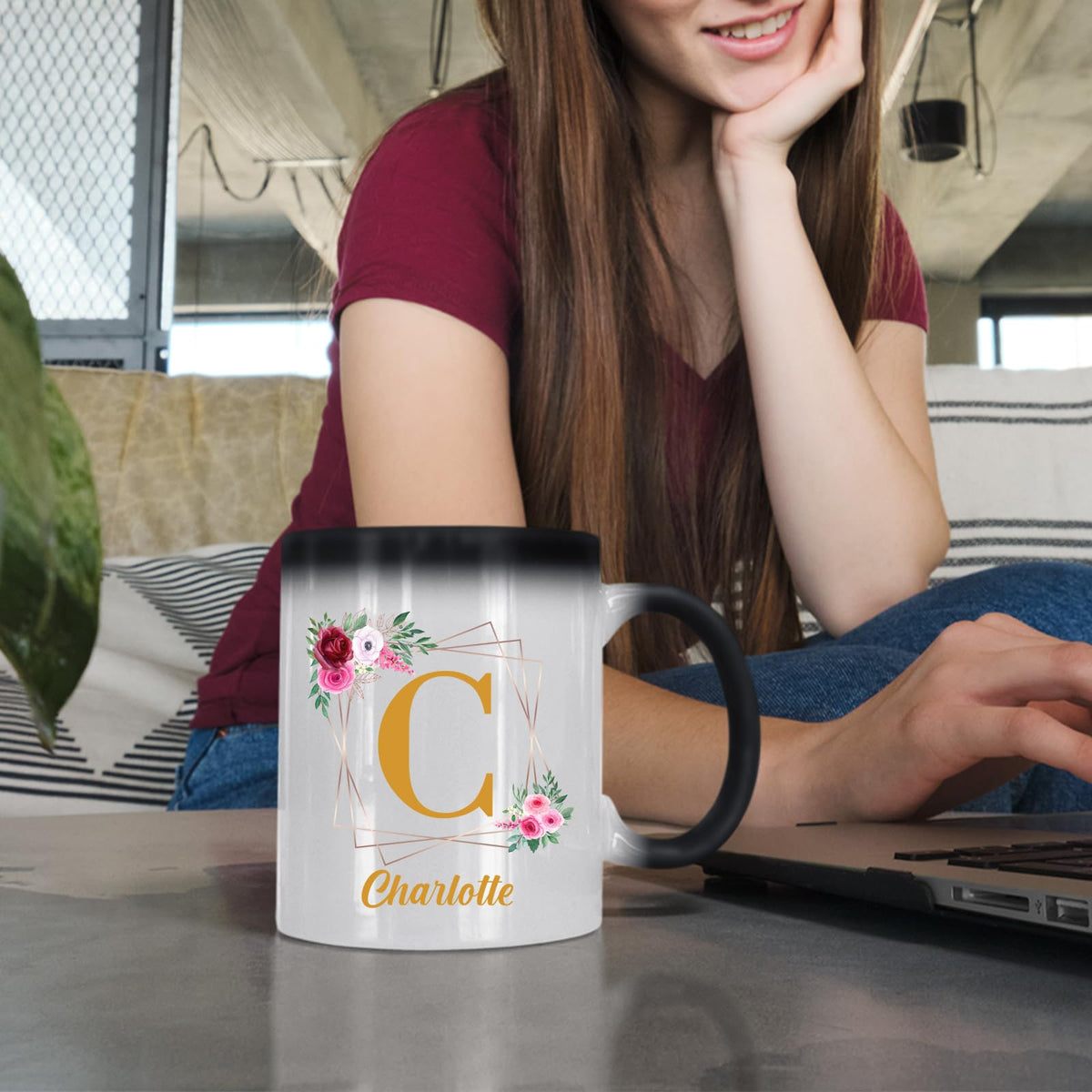 Woman at a laptop with a colour changing mug showing a gold initial in a floral frame and an example name