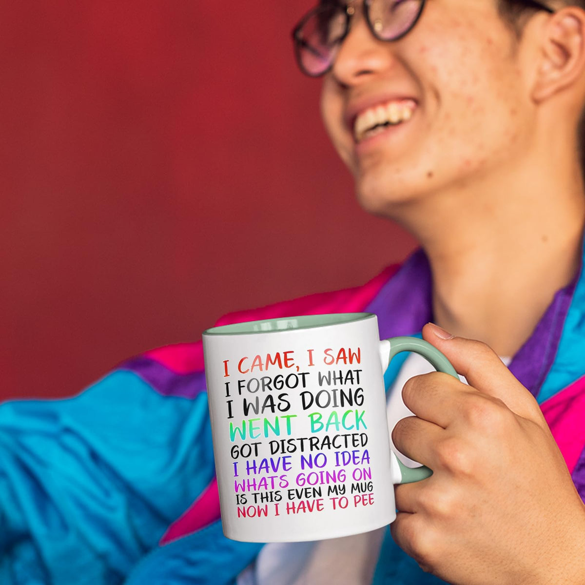 Smiling person in a colourful jacket holding the I Came I Saw I Forgot mug with a light green handle