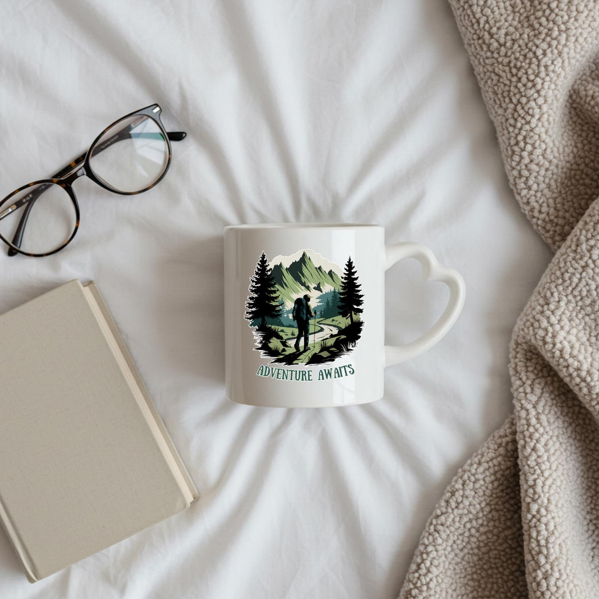 Adventure Awaits hiker heart-handle mug on a bed beside glasses and a book