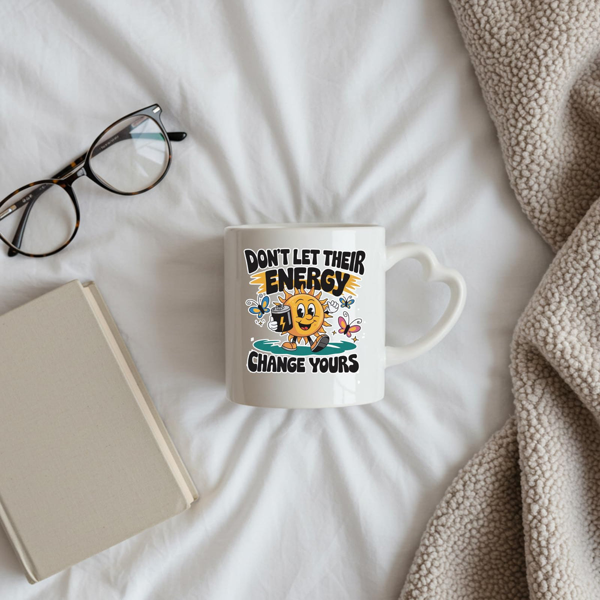 Positive Vibes sun heart-handle mug on a bed beside glasses and a book