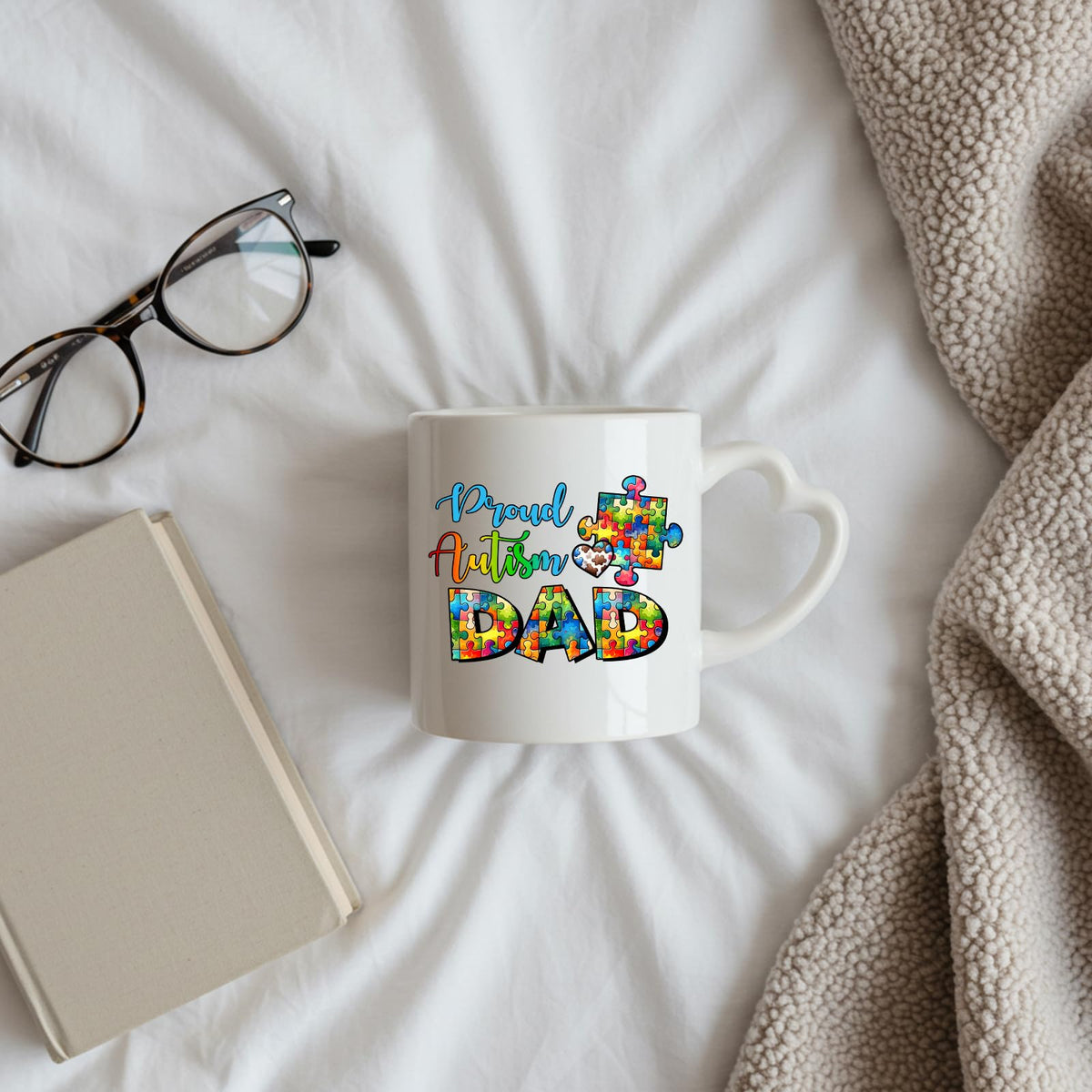 Proud Autism Dad heart-handle mug on a bed beside glasses and a book