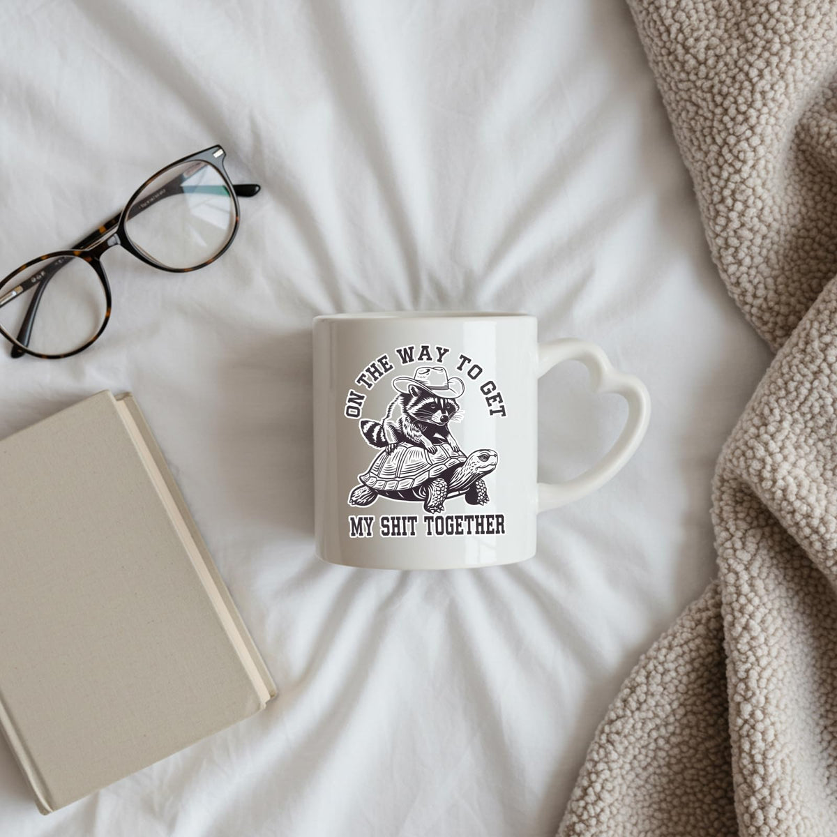 raccoon and tortoise heart-handle mug on a bed beside glasses and a book